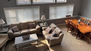Ein Wohnzimmer mit einem beigefarbenen Ledersofa, einem Holzesstzimmer mit Stühlen und großen Fenstern mit Jalousien.