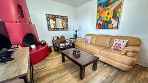 A living room with a red accent wall, a beige sofa, a wooden coffee table, a patterned armchair, and a colorful abstract painting. - Casita D - 2 Bed, 2 Bath (Single Level) (Santa Fe)