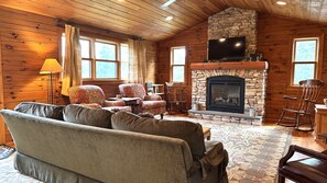 A log cabin interior with a stone fireplace, wooden walls, a sofa, and a television. - Overlook Cabin: E. Bluff Drive, Lake View, Hot Tub, Firepit, Central-AC, Private (Penn Yan)