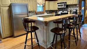 A kitchen with wooden cabinets, a stainless steel refrigerator, a microwave, and a bar stool area. - Overlook Cabin: E. Bluff Drive, Lake View, Hot Tub, Firepit, Central-AC, Private (Penn Yan)