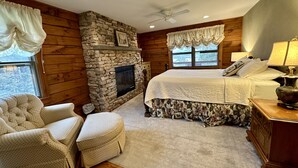 A cozy log cabin bedroom with a stone fireplace, a large bed with floral bedding, a wooden chair, and a nightstand with a lamp. - Overlook Cabin: E. Bluff Drive, Lake View, Hot Tub, Firepit, Central-AC, Private (Penn Yan)