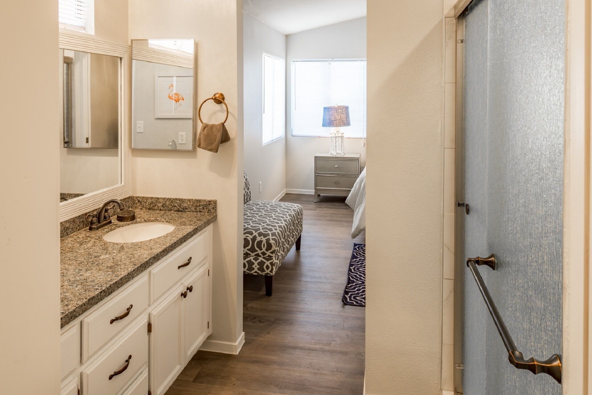 A narrow hallway leading to a bedroom with a patterned bedspread, a bedside table with a lamp, and a window with blinds.
