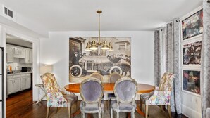 A dining area with a wooden table and chairs, a vintage painting on the wall, and a kitchen visible to the left.