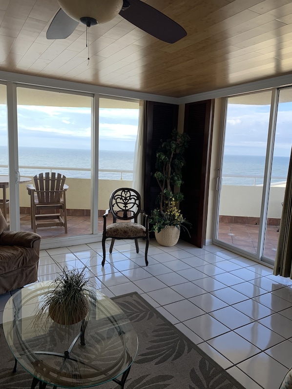 A spacious room with a large sliding glass door leading to a balcony with a view of the ocean. There is a chair, a table with a glass top, and a potted plant.