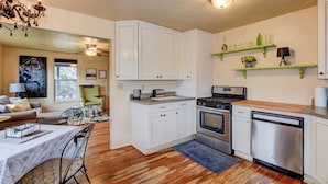 A compact kitchen with white cabinets, a stove, oven, and dishwasher. There is a dining area with a table and chairs, and a chandelier. - Abba House Downtown  (Prescott)