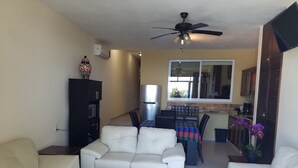 Una sala de estar con un sofá blanco, una mesa de centro, un comedor con una mesa y sillas, una cocina con un refrigerador y un ventanal, y un ventilador de techo.
