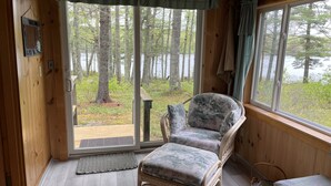 L’intérieur d’un chalet en bois, avec un fauteuil et un pouf près d’une grande fenêtre, une porte donnant sur l’extérieur et une petite table.