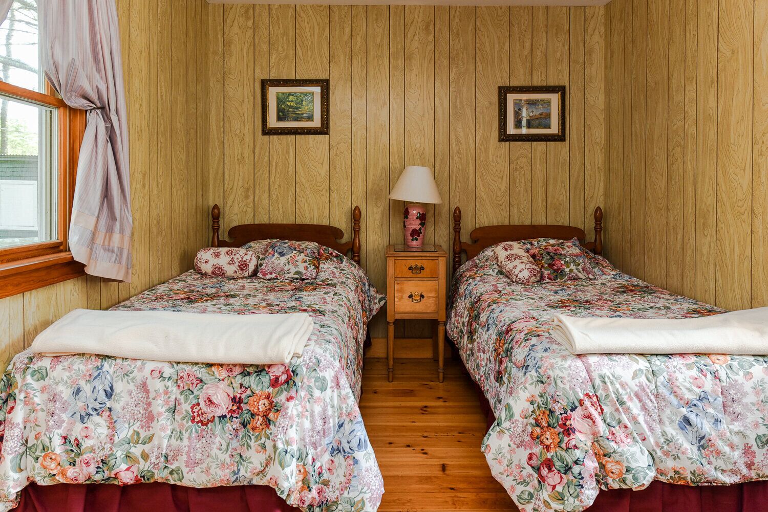 Une chambre avec deux lits en bois, chacun agrémenté d’une literie à motifs floraux et d’une table de chevet en bois avec une lampe.