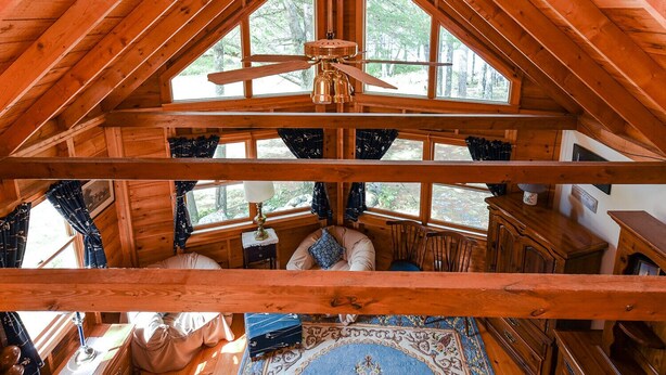 Une cabane avec un plafond haut, des poutres en bois, un ventilateur de plafond, une fenêtre avec des rideaux, un canapé, une table avec une lampe et un tapis.