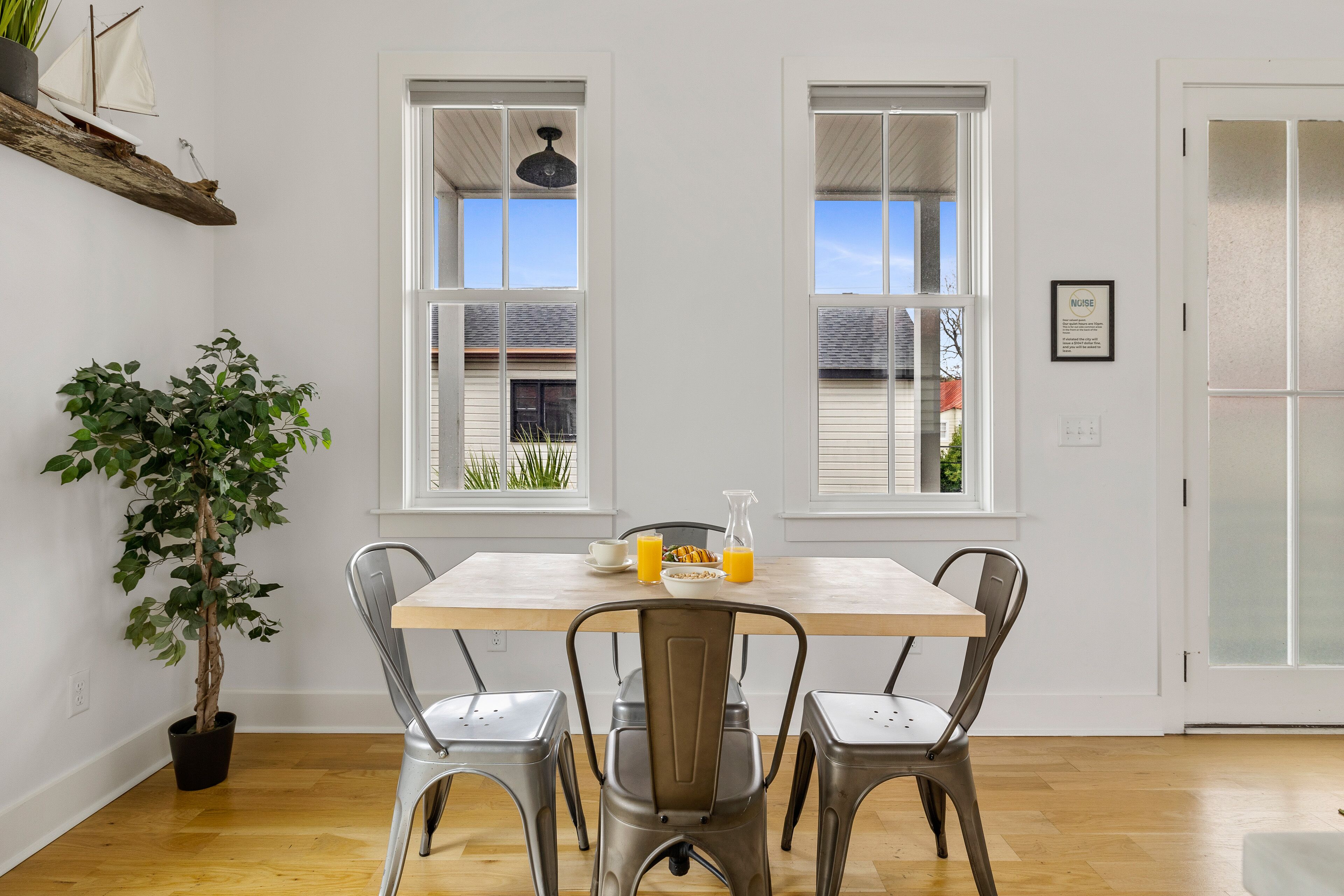 A dining area with a wooden table, four metal chairs, a potted plant, and two windows with blinds.