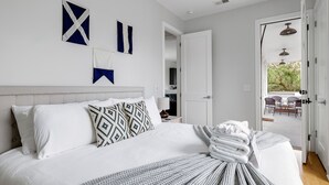 A neatly made bed with white bedding and a patterned throw, a white headboard, and a view of an outdoor patio through an open door. - Charming Cozy Porch! | Globetrotter Suite D (Charleston)