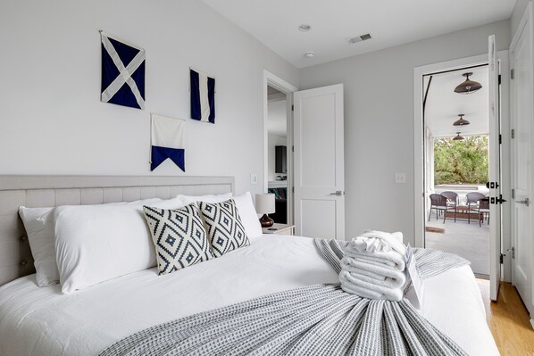 A neatly made bed with white bedding and a patterned throw, a white headboard, and a view of an outdoor patio through an open door. - Charming Cozy Porch! | Globetrotter Suite D (Charleston)
