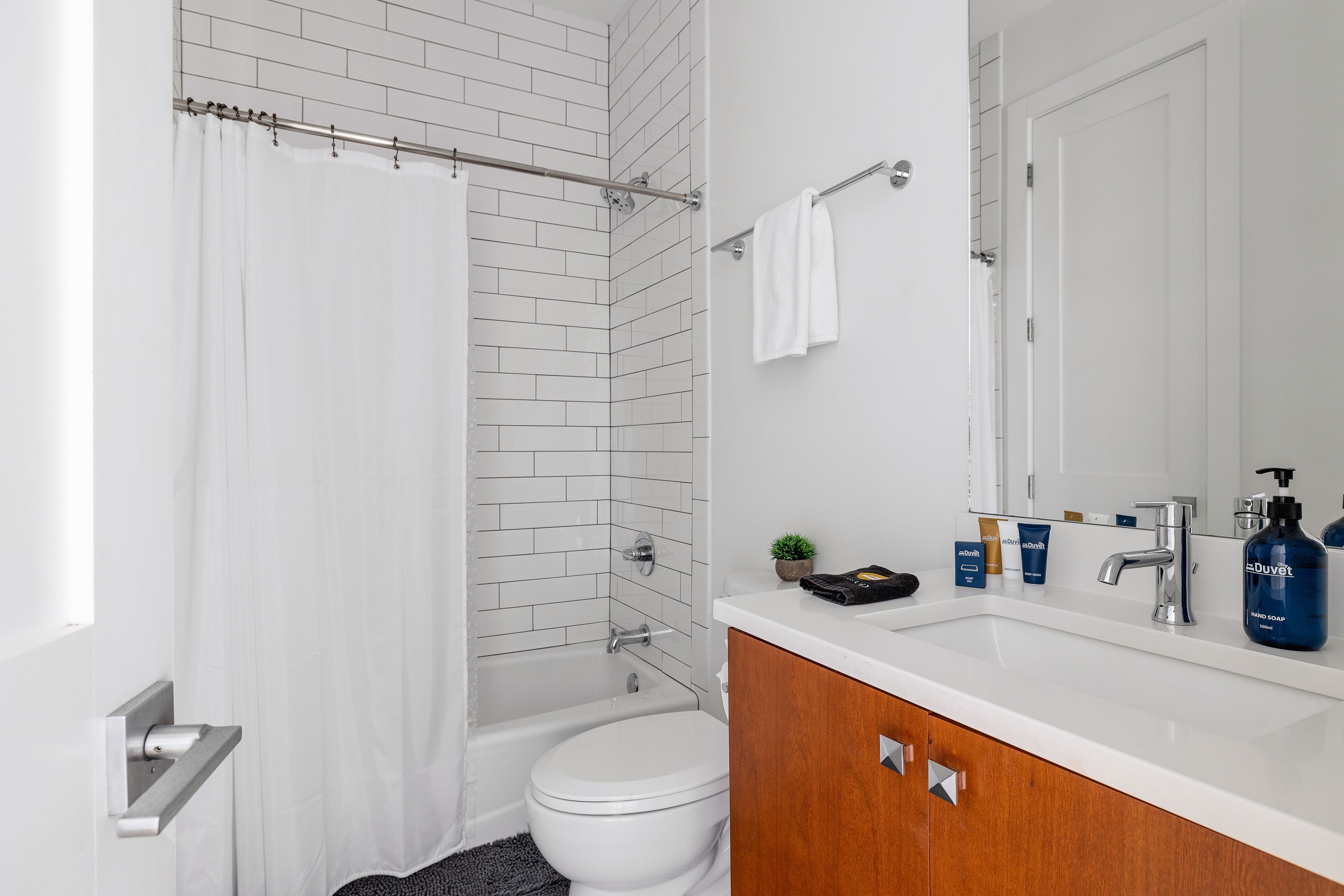 Une salle de bain moderne avec une douche carrelée de blanc, des toilettes blanches, un meuble-lavabo en bois avec un miroir et des articles de toilette posés sur le plan de travail.