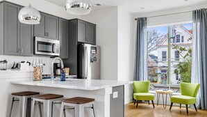 A modern kitchen with grey cabinets, stainless steel appliances, and a large island with bar stools. There are two green chairs and a small round table near a large window with a view of a house. - Charming Cozy Porch! | Globetrotter Suite D (Charleston)