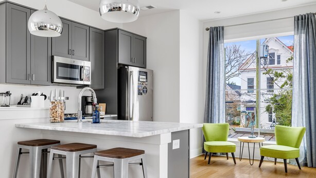 A modern kitchen with grey cabinets, stainless steel appliances, and a large island with bar stools. There are two green chairs and a small round table near a large window with a view of a house.