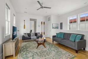 A spacious living room with a large grey sofa, a coffee table, and a TV on a wooden stand. There are two windows with white shutters, a ceiling fan, and a patterned rug on the floor.