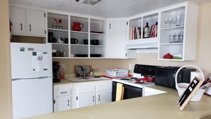 Une cuisine avec des meubles blancs, un réfrigérateur, une cuisinière et un four à micro-ondes. On trouve divers ustensiles de cuisine sur les plans de travail, notamment une bouilloire, une machine à café et un panier.