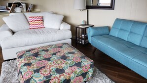 Un salon avec un canapé blanc, un canapé bleu, un pouf à motifs, une petite table avec une lampe et une bibliothèque.