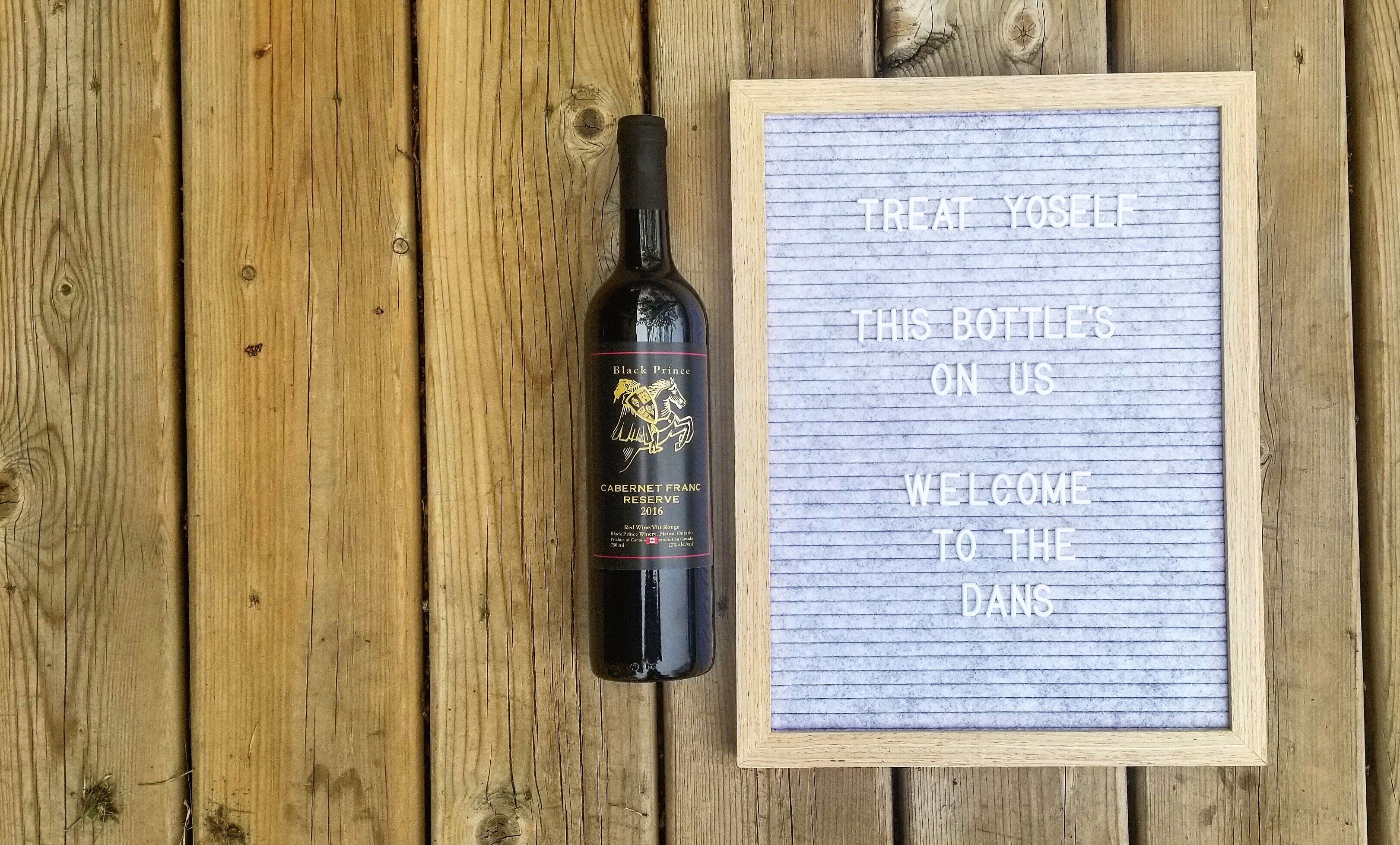 Une bouteille de vin Black Prince Cabernet Franc Reserve, millésime 2016, est posée à côté d’une pancarte encadrée portant un message d’accueil.