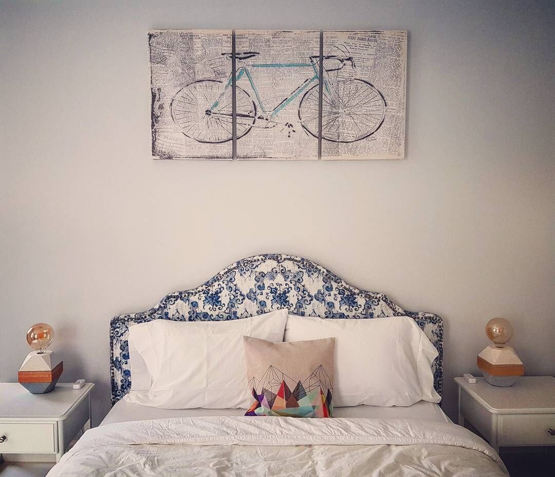 Une chambre à coucher avec un lit, du linge de lit blanc, une tête de lit ornée d’un motif floral bleu, une œuvre d’art représentant un vélo accrochée au mur, et deux tables de chevet surmontées de sphères décoratives.