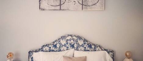 Une chambre à coucher avec un lit, du linge de lit blanc, une tête de lit ornée d’un motif floral bleu, une œuvre d’art représentant un vélo accrochée au mur, et deux tables de chevet surmontées de sphères décoratives.