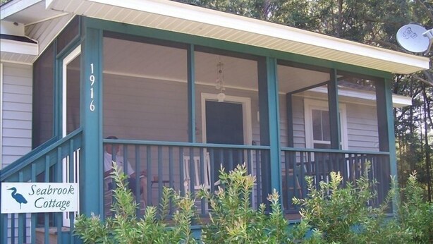 Una cabaña de dos pisos con un porche, un letrero que dice "Seabrook Cottage" y una antena parabólica en el techo.