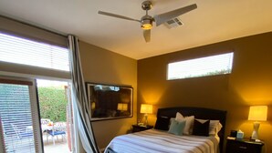 Une chambre à coucher avec un lit, des tables de chevet, un ventilateur de plafond et une vue sur l’extérieur, accessible par une porte-fenêtre coulissante.