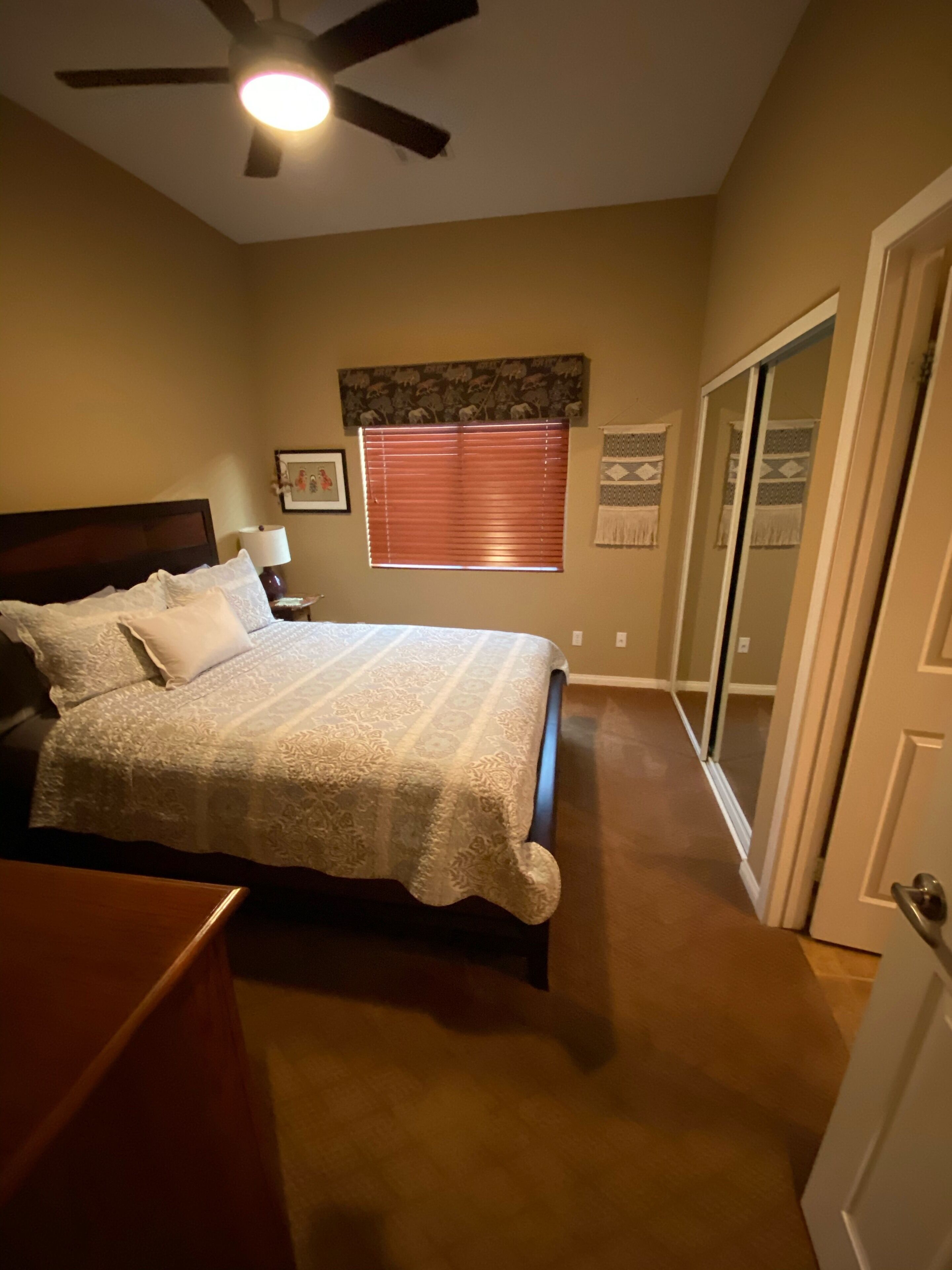 A bedroom with a bed, a nightstand, a ceiling fan, a window with blinds, a door, and a wall-mounted artwork.