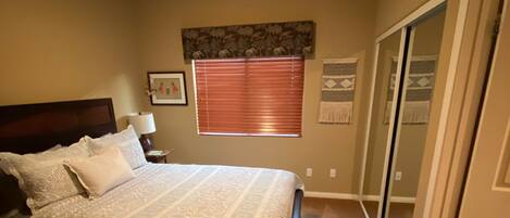 A bedroom with a bed, a nightstand, a ceiling fan, a window with blinds, a door, and a wall-mounted artwork.