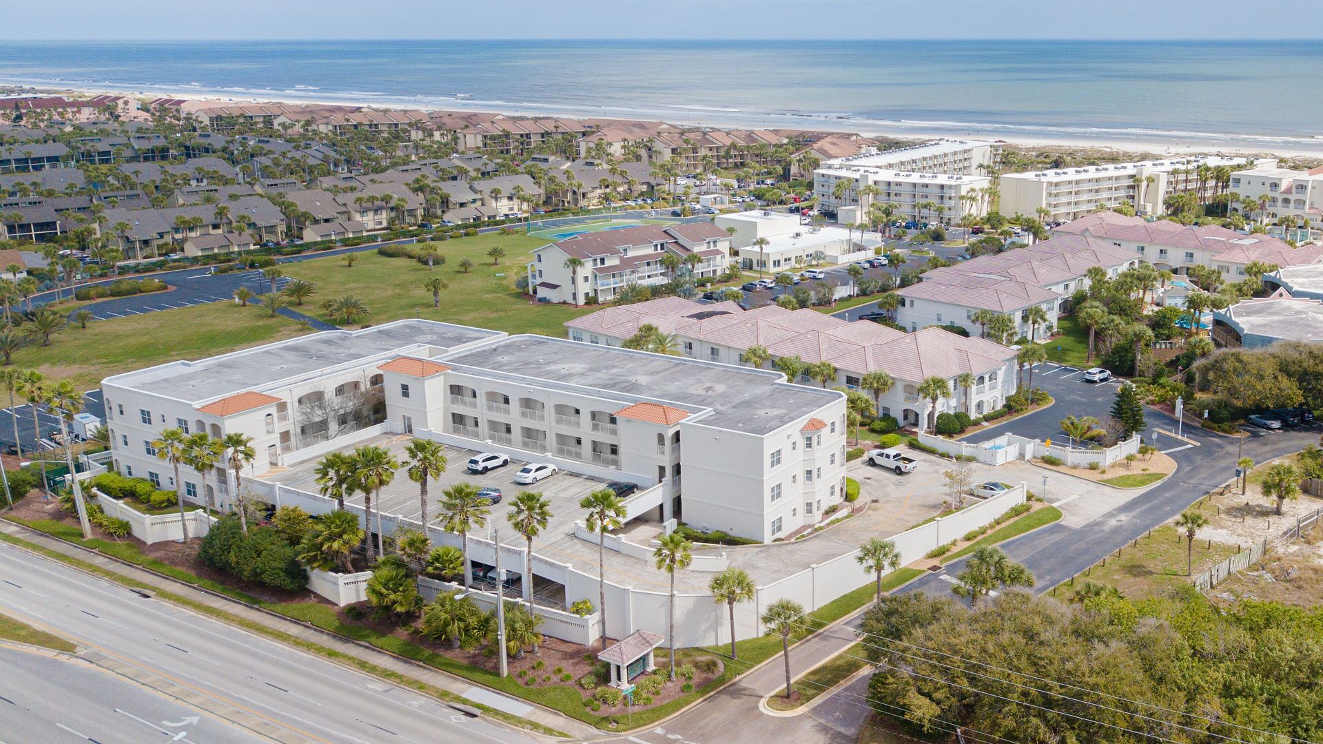A coastal residential area with a large building featuring multiple floors and a flat roof, surrounded by palm trees and a parking lot.