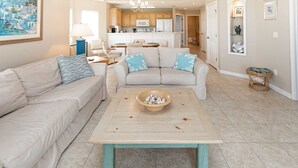 Ein Wohnzimmer mit einem beigefarbenen Sofa, einem Couchtisch mit einer Schale Muscheln und einer Küche im Hintergrund.