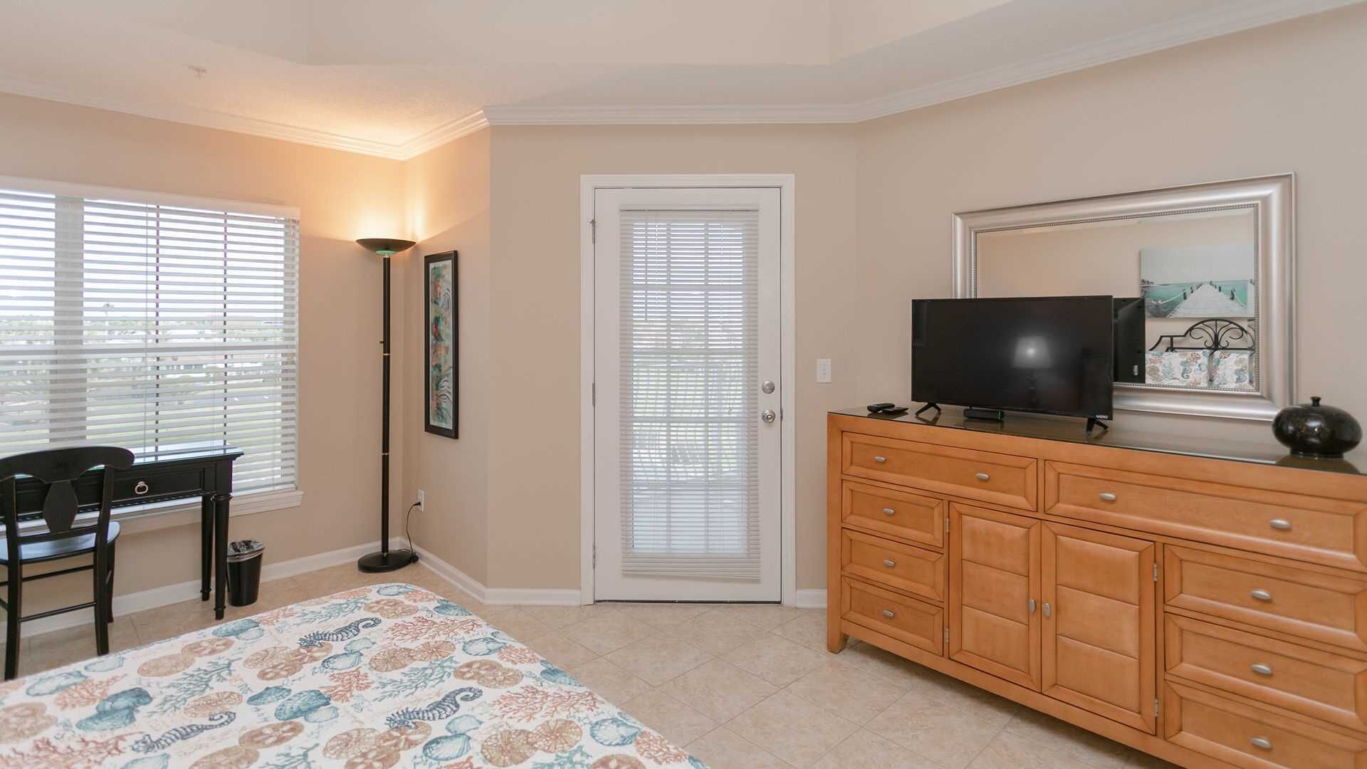 Une chambre à coucher comprenant un lit, un bureau, une chaise, une commode, un téléviseur et une porte avec des stores.
