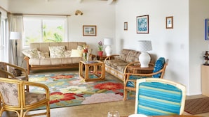 Ein Wohnzimmer mit einer Couch, Sesseln, einem Esstisch gedeckt mit Früchten und Gläsern sowie einem bunten Teppich.