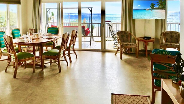 Ein geräumiges Zimmer mit einem großen Esstisch, gedeckt für sechs Personen, umgeben von Korbstühlen mit grünen Kissen. Ein Fernseher ist an der Wand befestigt, und das Zimmer verfügt über große Fenster mit Blick auf den Strand.