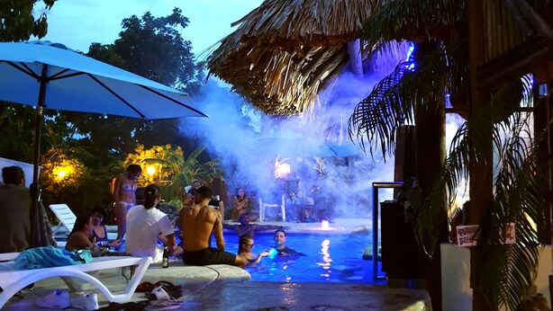 Ein Poolbereich mit Liegestühlen, einer Struktur mit Reetdach und einem Wasserspiel aus Stein.