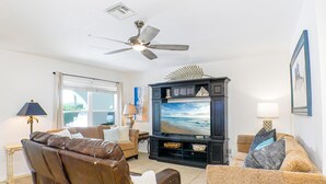 A living room with a large flat-screen TV, leather sofas, a ceiling fan, and a window with a view of the outdoors. - Hot Tub, Salt Air & Sunsets Your Naples Beach Retreat (Naples)