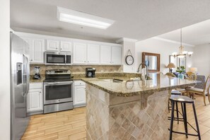 A modern kitchen with white cabinets, stainless steel appliances, a granite countertop, and a bar area with stools. - Spanish Key 504, next to Flora-Bama, beachfront (Pensacola)