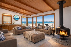 A cozy living room with a fireplace, large windows overlooking the beach, and a wooden ceiling. - Sandy BEACHFRONT Surfside - at Corcoran Beach, Newly Remodeled w/ HOT TUB! (Santa Cruz)