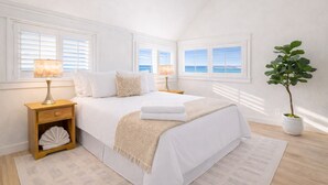 Ein Schlafzimmer mit einem Bett, zwei Nachttischen, einer Stehlampe und einem Blumentopf, mit Blick aufs Meer durch die Fenster.
