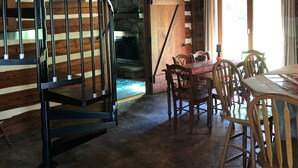 Un intérieur rustique avec un escalier en bois, une cheminée en pierre, un coin repas avec des chaises en bois et un ventilateur de plafond.