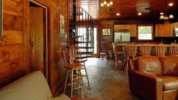 L’intérieur d’un chalet rustique est agrémenté d’un escalier en bois, d’un coin repas avec des chaises en bois, d’un canapé en cuir et d’une cuisine avec des placards en bois.