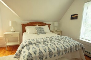 A bedroom with a wooden headboard, a patterned bedspread, two bedside tables with lamps, a small painting on the wall, and a window with blinds. - Your Kennebunkport Home Base (Kennebunkport)
