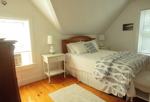 A bedroom with a sloped ceiling, a wooden headboard, two bedside tables with lamps, a bed with a patterned duvet, and a white rug. - Your Kennebunkport Home Base (Kennebunkport)