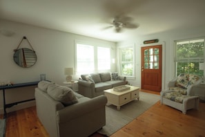 Ein Wohnzimmer mit einer Couch, einem Couchtisch, einem Beistelltisch und einem Sessel. Eine Tür mit Fenster, ein Deckenventilator und ein wandmontierter Spiegel sind ebenfalls vorhanden.