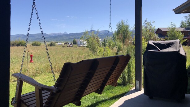 A wooden swing chair under a covered porch with a view of a grassy field and mountains in the background. - Your Basecamp In the Tetons.  (Victor)