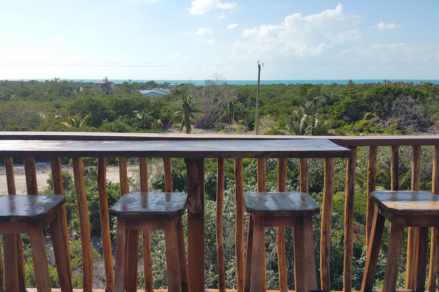 A wooden deck with three stools and a table, overlooking a coastal landscape with dense vegetation and a view of the ocean.