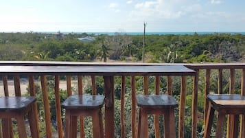 A wooden deck with three stools and a table, overlooking a coastal landscape with dense vegetation and a view of the ocean.