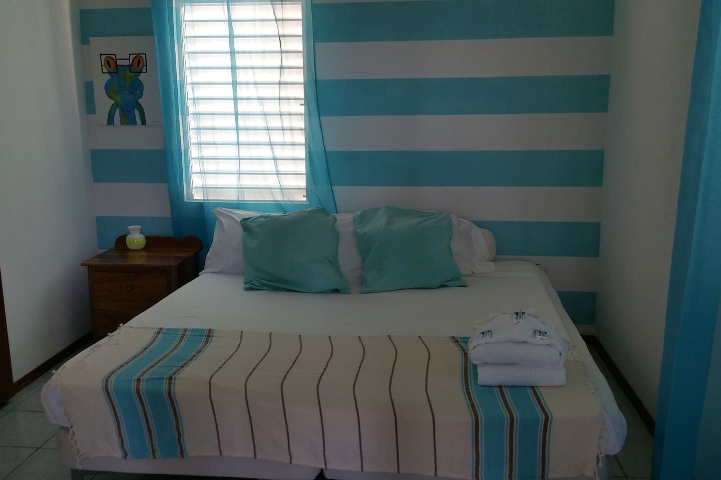 Une chambre à coucher avec un lit, des oreillers, une table de chevet, une fenêtre avec des rideaux et un mur à rayures horizontales.