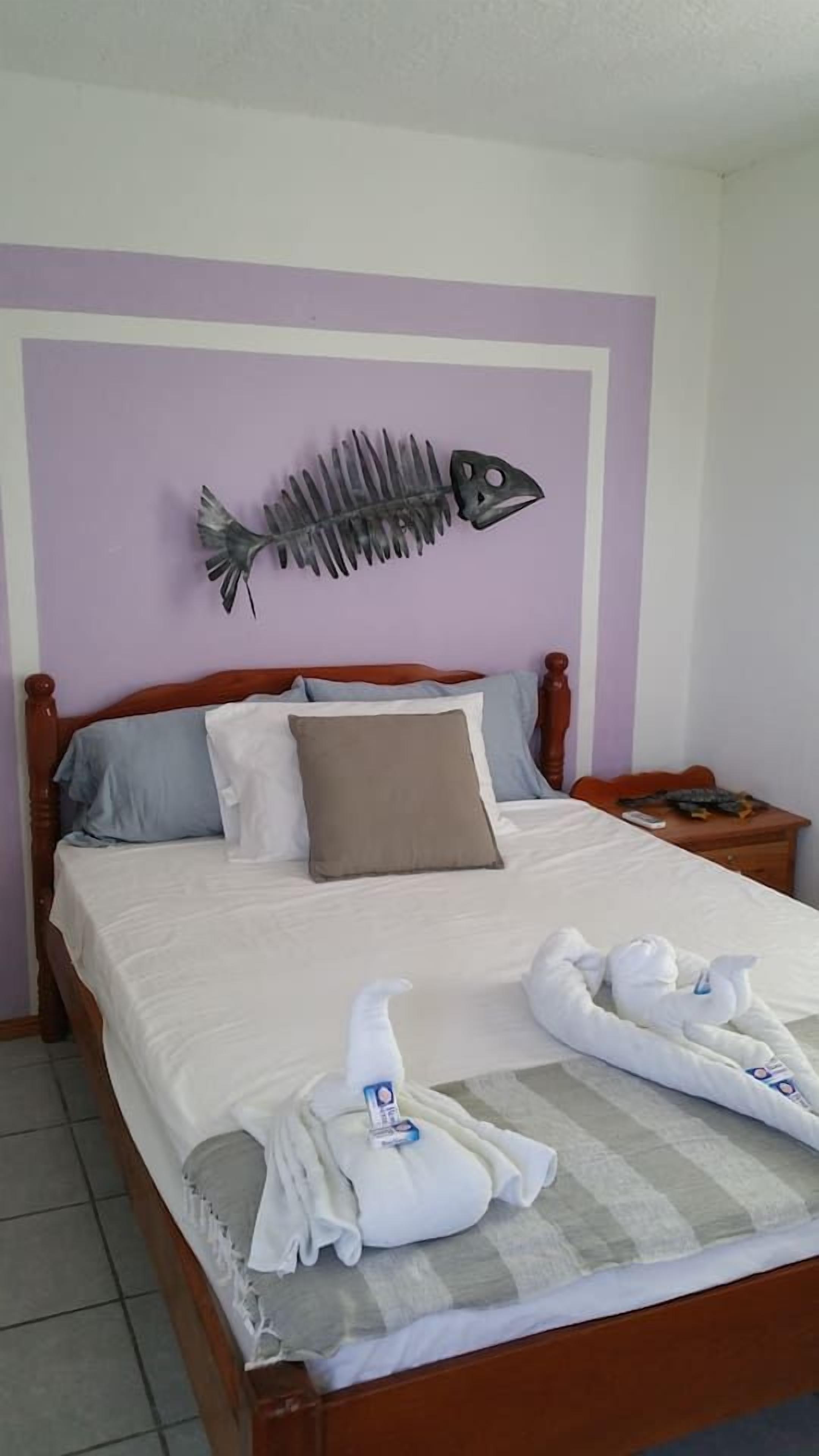Un letto ben rifatto con cuscini, una testiera in legno, una decorazione a forma di pesce appesa al muro e un comodino con un telecomando.