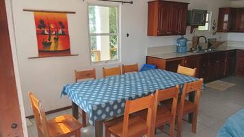 A dining area with a table set for six, wooden chairs, a blue tablecloth, and a view of the kitchen with cabinets and a water dispenser.